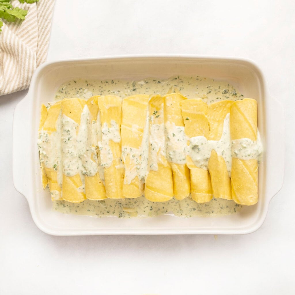 White Chicken Enchiladas rolled in baking dish with sauce on top.