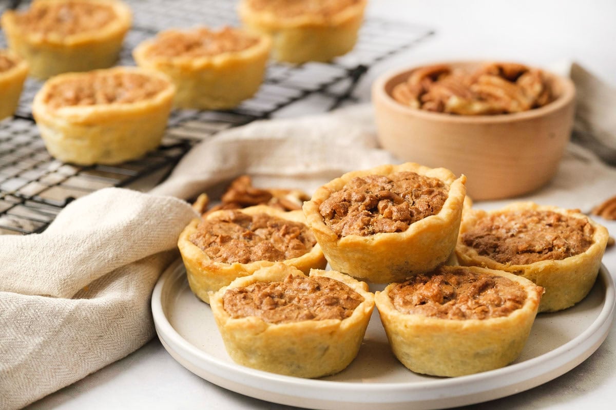 Mini Pecan Pies on plate and cooling rack with bowl of pecans in background.